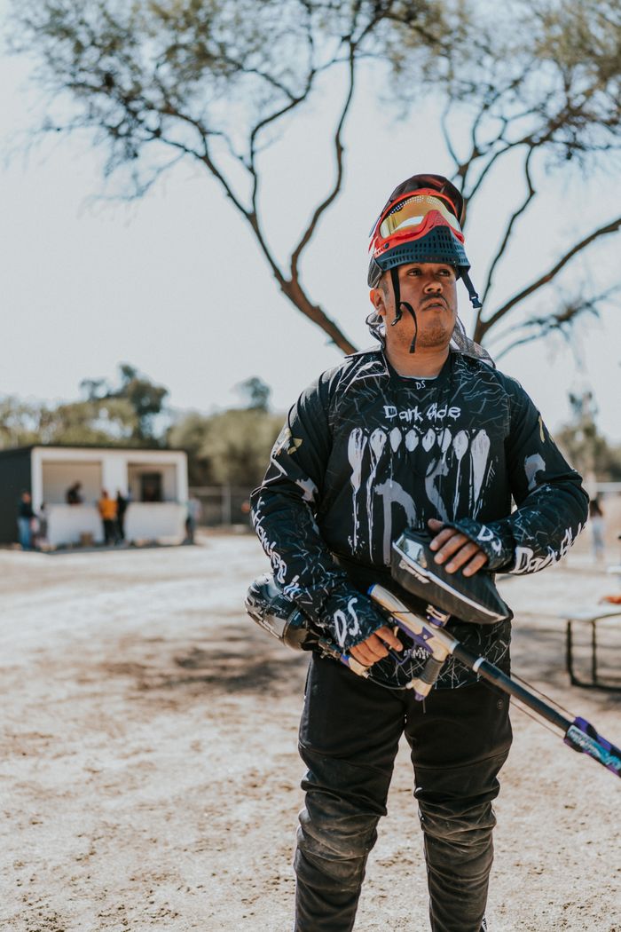 A paintball player stands outdoors in full gear holding a paintball gun, ready for action.