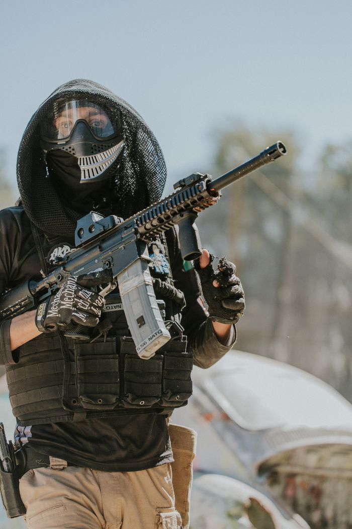A paintball player dressed in tactical gear and mask holding a paintball rifle outdoors.